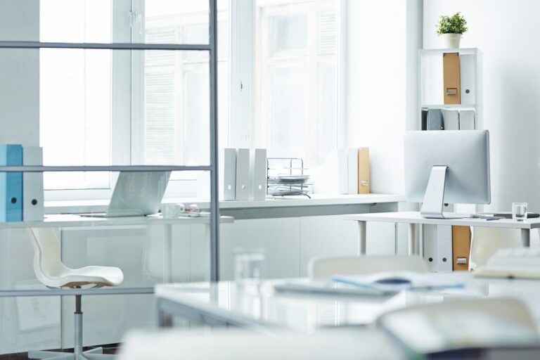 Ein helles, minimalistisches Büro mit weißen Möbeln, einem Computerbildschirm und Aktenordnern auf Regalen. Große Fenster lassen viel Tageslicht herein.